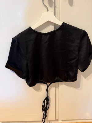 Svart croppad satintopp med snörning - Snygg svart croppad topp i satin med korta ärmar och snörning i midjan. Perfekt för dig som gillar trendiga och stilrena plagg med en lyxig känsla. Toppen har en enkel rund halsringning och glansigt material.