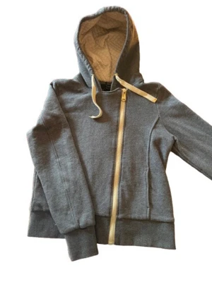 Goldberg - grå hoodie med sned dragkedja - Snygg grå hoodie från The Goldbergs med sned dragkedja och quiltad insida i huvan. Tryck på insidan med texten 'The Goldberg Standard'. Mjukt material och ribbade muddar vid ärmar och midja. Perfekt för en chill och avslappnad stil.