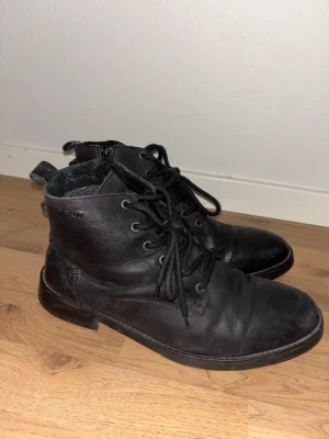 Svarta snörade läderkängor - Säljer ett par svarta kängor i läder med snörning och dragkedja på sidan. De har rundad tå och robust sula, samt detaljer i metall vid snörningen. Insidan verkar vara fodrad för extra komfort. Perfekta för dig som vill ha en stilren och klassisk look.