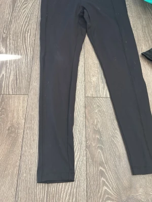 Svarta leggings med vit logga - Säljer ett par svarta leggings med en vit logga vid höften. De har en tight passform och är tillverkade i ett stretchigt material som sitter skönt på kroppen. Perfekta till träning eller chill. 