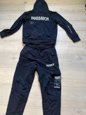 Svart Hoodrich loungewear set - Snyggt svart loungewear set från Hoodrich med hoodie och matchande byxor. Hoodien har tryckt logga framtill och på ärmen, medan byxorna har logga och grafiskt tryck på benet samt praktisk ficka. Perfekt för chill dagar hemma eller streetwear vibes.