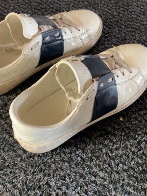 Vita sneakers med svart detalj - Snygga vita sneakers från Valentino Garavani med bred svart läderdetalj över snörningen. Skorna har klassisk låg profil, vita snören och diskret logga på plösen. Perfekta för dig som gillar stilrena och exklusiva sneakers.