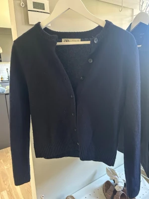 Mörkblå cardigan från Zara - Mysig mörkblå cardigan från Zara med rund halsringning och knäppning framtill. Tröjan har långa ärmar och ribbade muddar vid ärmslut och nederkant. Perfekt att slänga över en topp för en enkel och stilren look.