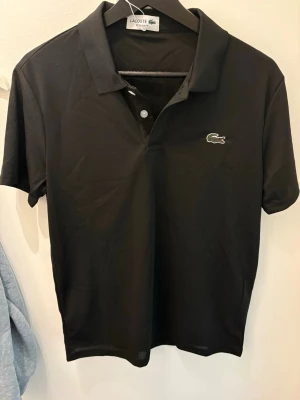 Svart pikétröja från Lacoste - Klassisk svart pikétröja från Lacoste med korta ärmar och krage. Tröjan har knappslå framtill och den ikoniska krokodilloggan broderad på bröstet. Perfekt till både jeans och shorts för en clean look.