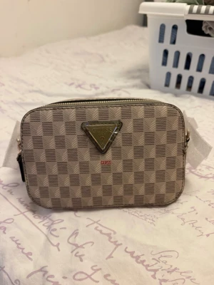 Beige-rutig handväska från Guess - Snygg beige handväska från Guess med rutigt mönster och guldfärgade detaljer. Väskan har en rektangulär form, dragkedja upptill och den klassiska Guess-loggan i metall framtill. Perfekt för att bära det viktigaste med stil.