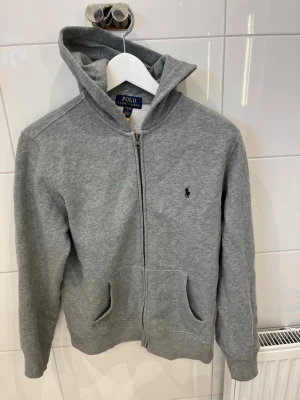 Grå hoodie från Polo Ralph Lauren - Snygg grå hoodie från Polo Ralph Lauren med dragkedja framtill och klassisk blå logga broderad på bröstet. Hoodien har huva, två fickor och är gjord i mjukt sweatshirtmaterial. Perfekt för en chill och avslappnad stil. Storlek 14-16 så den är för barn 
