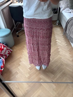 Lång röd mönstrad kjol - Säljer en lång kjol i rött och vitt med ett snyggt mönster. Kjolen har resår i midjan för skön passform och är gjord i ett lätt och luftigt material, perfekt för varmare dagar. Passar till både sneakers och sandaler.
