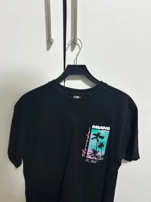 Svart Miami t-shirt med tryck. Storlek S - Cool svart t-shirt med färgglatt Miami-tryck i turkos, rosa och vitt både fram och bak. Motiv med palmer och strandvibbar. Klassisk rund hals och korta ärmar. Perfekt för dig som gillar streetstyle och grafiska prints. 