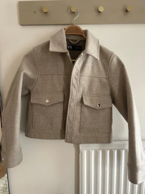 Beige höst jacka med fickor - Stilren beige höst jacka  i ullmix med två stora bröstfickor och silverfärgade knappar. Rak passform, klassisk krage och långa ärmar med knapp vid ärmslut. Perfekt lager-på-lager-plagg för kyligare dagar.