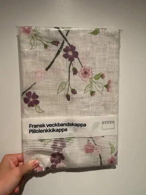 Fransk veckbandskappa med blommor - Säljer en fransk veckbandskappa från Hemtex i vitt tyg med lila och rosa blommor samt gröna blad. Gardinen har dold upphängning och är tillverkad av polyester och bomull. Perfekt för att ge rummet en fräsch och vårig känsla.