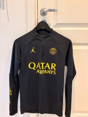 Svart PSG träningströja med gula detaljer - Snygg svart långärmad träningströja från PSG x Jordan med gula detaljer och loggor. Halvlång dragkedja framtill och tryckta Qatar Airways och Paris Saint-Germain-emblem på bröstet. Perfekt för fotbollsträning eller chill.