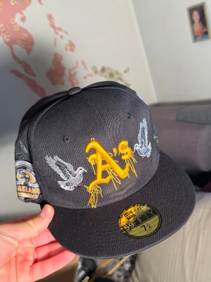 Svart Oakland A's snapback keps - Snygg svart snapback keps med gult Oakland A's broderi framtill, detaljer med duvor och coola skelett- och blomsterpatchar på sidorna. Platt skärm och klassisk 59FIFTY-stil. Perfekt för dig som vill sticka ut med unik design.