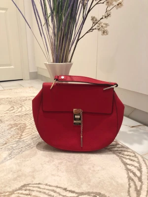 Röd Chloé handväska  - Säljer en snygg röd handväska från Chloé - det är inte det riktiga märket. Väskan har en rund form, lock med guldfärgat lås och kedjedetalj framtill. Kort axelrem med metallfästen ger en exklusiv känsla. Perfekt accessoar för att lyfta din outfit.