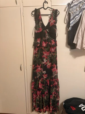Blommig långklänning från Guess - Snygg långklänning från Guess med svart bas och färgglatt blommönster i rosa, rött och grönt. Klänningen har v-ringning, volangdetaljer och är ärmlös. Perfekt för sommarens alla tillfällen!