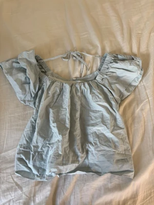 Ljusblå offshoulder topp med knyt - Supersöt ljusblå topp med offshoulder-modell och korta, vida ärmar. Toppen har knytband i nacken och fina veckade detaljer längst ner. Perfekt för sommarens alla tillfällen!