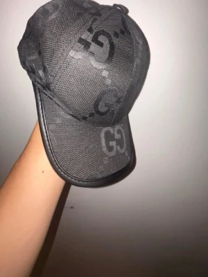 Svart keps med GG-mönster - Snygg svart keps med tydligt GG-mönster över hela ytan. Kepsen har böjd skärm och är tillverkad i ett slitstarkt tygmaterial. Perfekt för dig som gillar stilrena accessoarer med en exklusiv touch.