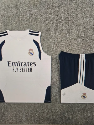 Real Madrid matchställ och röd tröja - Säljer ett Real Madrid matchställ med vit ärmlös tröja och mörkblå shorts med klubbmärke och sponsortryck. Ingår även en röd kortärmad Real Madrid-tröja med vitt tryck och klubbmärke. Perfekt för fotbollsträning eller match.