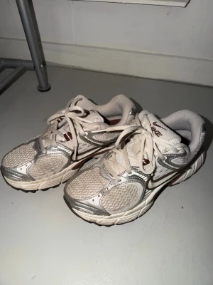 Nike vita och beige sneakers - Säljer ett par vita och beige Nike sneakers med silvriga detaljer och meshpaneler för extra ventilation. Skorna har klassisk snörning och en chunky sula som ger skön komfort. Perfekta för dig som gillar sportig och trendig stil.
