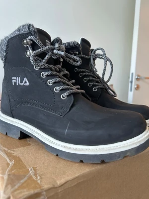 Svarta kängor från FILA med foder - Säljer ett par svarta kängor från FILA med grov sula, gråmelerade skosnören och mjukt grått foder vid skaftet. Kängorna har metallhål för snörningen och en vit kontrastsula. Perfekta för kalla dagar och riktigt snygga till jeans.