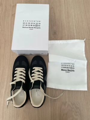 Maison Margiela gats - Maison Margiela sneakers i svart läder och mocka med beigea skosnören och gummisula. Klassisk low-top modell med stilren design och diskreta detaljer. Kommer med originalkartong och dustbag. Perfekta för dig som gillar minimalistisk och trendig stil.
