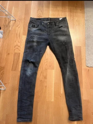  Elias Rumelis Jeans - Säljer ett par svarta slim jeans från Elias Rumelis med slitningar och tvättade detaljer. Jeansen har fem fickor, låg midja och knappgylf. Snygg streetstil och perfekt passform för dig som gillar en smal siluett.
