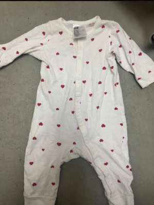 Vit pyjamas med röda hjärtan H&M - Supersöt vit body från H&M i storlek 62, dekorerad med små röda hjärtan över hela plagget. Långärmad modell med tryckknappar framtill och i grenen för enkel påklädning. Tillverkad i mjuk bomull som känns skön mot huden.