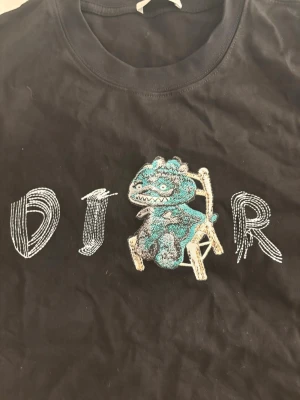 Svart t-shirt med broderad dinosaurie - Cool svart t-shirt med ett broderat motiv av en blå dinosaurie som sitter på en stol och texten 'DJAR' på bröstet. Rund hals och korta ärmar. Perfekt för dig som gillar unika och lekfulla prints.