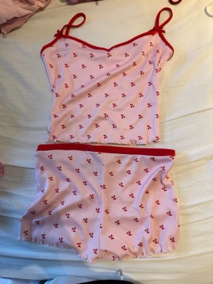 Rosa pyjamas-set med körsbärsmönster - Sött pyjamas-set med linne och shorts i ljusrosa med små röda körsbär och röda detaljer. Linnet har tunna axelband och rosetter vid axlarna, medan shortsen har en röd resår i midjan. Perfekt för mysiga kvällar hemma.