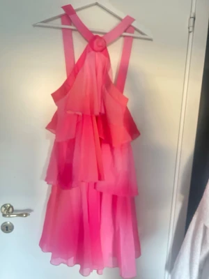 Rosa volangklänning med axelband - Säljer en superfin rosa kortklänning med volanger och breda axelband som korsas i ryggen. Klänningen har flera lager i ett lätt, flowy material och en snygg omlottkänsla. Perfekt för dig som vill sticka ut med färg och volym! Aldrig använd! Prislappar kvar 