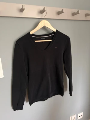 Svart V-ringad tröja Tommy Hilfiger - Klassisk svart V-ringad damtröja från Tommy Hilfiger med diskret logga på bröstet. Tillverkad i mjuk bomull, långärmad och med ribbade muddar vid ärmslut och nederkant. Perfekt för en stilren och enkel look.