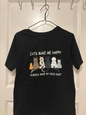 Svart t-shirt med kattmotiv - Svart t-shirt med tryck av flera katter i olika färger och texten 'Cats make me happy, humans make my head hurt' på framsidan. Klassisk rund hals och korta ärmar. Perfekt för dig som älskar katter och vill ha en rolig statement-tee.