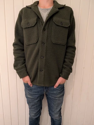 Olivgrön fleece overshirt från Polo Ralph Lauren - Säljer en olivgrön overshirt i mjuk teddyfleece från Polo Ralph Lauren. Skjortan har två stora bröstfickor med lock och knappar framtill. Klassisk krage och diskret broderad logga vid nederkanten. Perfekt för lager på lager och riktigt skön att ha på sig.