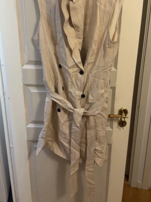 Beige ärmlös byxdress med bälte - Stilren beige ärmlös byxdress med tunna vertikala ränder och knappar framtill. Byxdressen har ett matchande tygbälte i midjan och en djup v-ringning. Perfekt för dig som gillar en chic och avslappnad look. Obs, den behöver styrkas annars är den helt ny. Med prislapp också