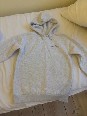 Grå hoodie med tryck 'Ma chérie' - Ljusgrå hoodie med huva och dragsnören. På bröstet står det 'Ma chérie' och på ryggen finns ett stort tryck med ett hjärta och pil samt texten 'Ma chérie'. Mjuk och skön, perfekt för chill dagar.