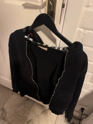 Svart Burberry hoodie med dragkedja - Säljer en nu min svarta burberry hoodie med dragkedja och huva. Insidan av huvan har ett rutigt mönster i svart, vitt och rött som ger en snygg kontrast. Hoodien har fickor framtill och ribbade muddar vid ärmar och nederkant. Perfekt för en chill och avslappnad stil. Storlek S den är lite liten i storleken så skulle rekommendera till folk mellan cirka (165-170). 100% äkta. Kontakta mig om frågor!
