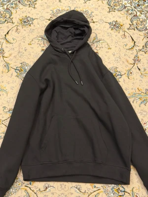 Svart hoodie med huva och ficka - En stilren svart hoodie med klassisk huva och dragsnören. Hoodien har en stor magficka framtill och ribbade muddar vid ärmar och nederkant. Perfekt för en avslappnad och trendig look. Tillverkad i mjukt material för extra komfort.