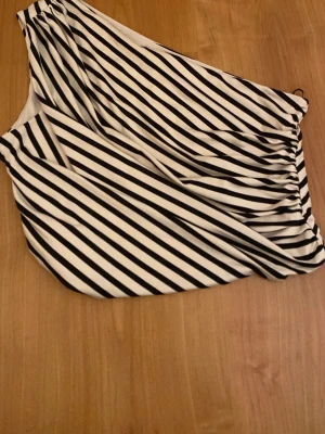 Randig oneshoulder topp - Snygg oneshoulder topp med svartvita diagonala ränder. Toppen är ärmlös och har en asymmetrisk skärning som ger en cool och modern look. Perfektion att styla med jeans eller kjol för en trendig outfit.