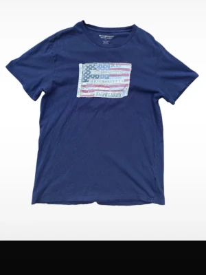 Blå t-shirt från Ralph Lauren - Snygg marinblå t-shirt från Ralph Lauren med amerikansk flagga och texten 'Denim & Supply' på bröstet. Klassisk rund hals och korta ärmar. Perfekt för en avslappnad och cool stil.