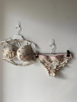 Blommigt bikini-set med volang - Söt bikini med blommigt mönster i pastellfärger. Överdelen har vadderade kupor och knyt i nacken för extra stöd. Underdelen har en rosa randig midja och volangkant. Perfekt för sommarens stranddagar! Överdelen är i storlek 70C och underdelen i 34. 