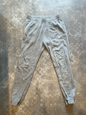 Gråa mjukisbyxor från Nike - Snygga gråa mjukisbyxor från Nike med klassisk logga broderad på vänster ben. Byxorna har resår i midjan, muddar vid bensluten och två sidofickor. Perfekta för chill eller träning, tillverkade i mjukt och bekvämt material.