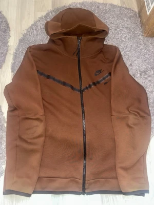 Brun Nike hoodie med dragkedja - ÄKTA.        Snygg brun hoodie från Nike med hel dragkedja framtill och svart Nike-logga på bröstet. Hoodien har en huva och ett svart diagonalt band över bröstet som detalj. Perfekt för en avslappnad och sportig stil. Obs fins en pytte lite fläck 
