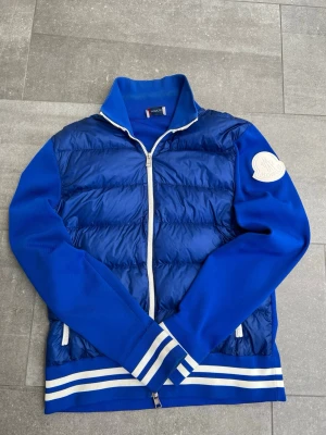 Blå cardiganjacka från Moncler - Snygg blå cardiganjacka från Moncler med quiltad front och stickade ärmar. Jackan har vit dragkedja, ribbade muddar med vita ränder och en stor Moncler-logga på ärmen. Perfekt för dig som vill ha en sportig och trendig look. Den är i mycket gott skick och knappt använd!