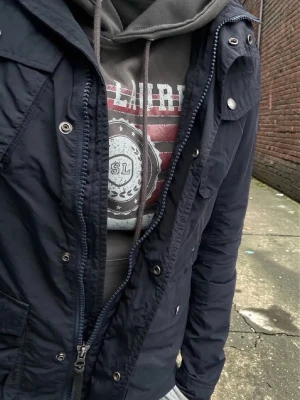 Grå Saint Laurent hoodie med tryck - Grå hoodie från Saint Laurent med stort tryck framtill i rött, vitt och grått. Trycket har texten 'SAINT LAURENT' och ett emblem med SL. Hoodien har huva med dragsko och en stor magficka. Perfekt för en avslappnad och cool stil.