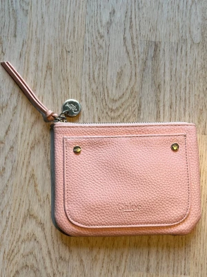 Ljusrosa plånbok från Chloé - Supersöt ljusrosa plånbok från Chloé i mjukt, strukturerat läder. Plånboken har en praktisk dragkedja, dekorativa guldfärgade nitar och en rund metallbricka med Chloé-logga. Perfekt för kort och småmynt, med stilren och trendig design. Tveka inte på att skriva en fråga eller be om fler bilder.🌸🌸