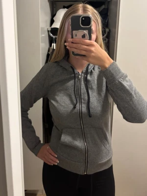 Grå zip hoodie med huva - Säljer en enkel och stilren grå hoodie med dragkedja framtill och justerbar huva med svarta snören. Hoodien har två fickor på magen och ribbade muddar vid ärmslut och nederkant. Perfekt för en avslappnad look.