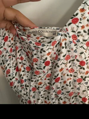Blommig långklänning från Mudo Collection - Långklänning med smala axelband och v-ringning från Mudo Collection. Klänningen är vit med ett blommigt mönster i rött, grönt och orange. Volangdetaljer längs kjolen ger en romantisk känsla. Perfekt för sommarens alla tillfällen! Är som en 38a 