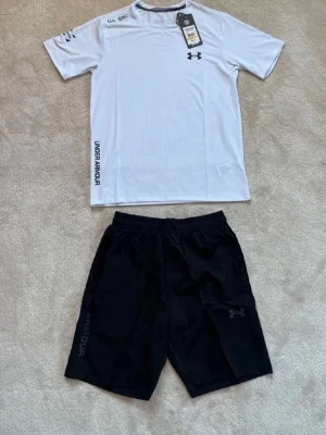 Under Armour träningsset svart/vit - Snyggt träningsset från Under Armour med en vit t-shirt och svarta shorts. T-shirten har tryckt logga på bröstet och text på ärm och sida. Shortsen har elastisk midja och diskret logga på benet. Perfekt för gym eller löpning.