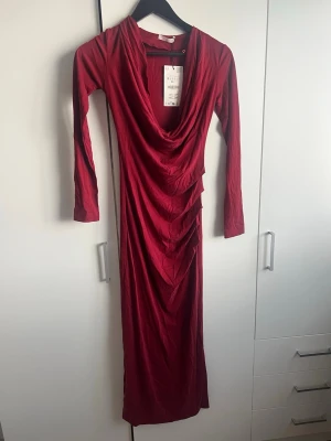 Lång vinröd klänning med drapering - Snygg vinröd långklänning med draperad framsida och djup urringning. Klänningen har långa ärmar och är tillverkad i ett mjukt, följsamt material som ger en elegant siluett. Perfekt för dig som vill sticka ut med stil.