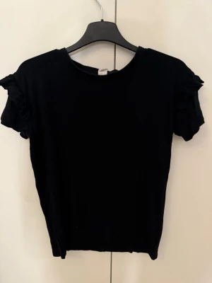Svart topp med volangärmar - Svart topp med korta ärmar och volangdetaljer vid axlarna. Toppen har en enkel, rak passform och är tillverkad i mjukt material som känns skönt mot huden. Perfekt att styla med jeans eller kjol.
