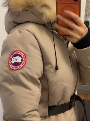 Beige jacka med pälshuva - Snygg beige jacka  med svart bälte i midjan och stor huva med fluffig päls. Jackan har en patch på ärmen och svarta muddar vid ärmsluten. Perfekt för kalla vinterdagar och riktigt varm. Pälsen är äkta.